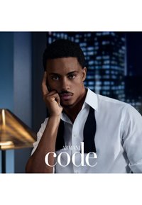 Homme en chemise blanche avec un nœud papillon noir défait tenant un téléphone à l'oreille, lumières de la ville visibles à travers la fenêtre derrière lui, texte Armani Code en surimpression.