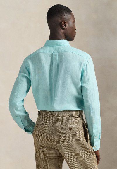 Camisa de lino azul claro con mangas largas, cuello puntiagudo y pliegue en la espalda, combinada con pantalones marrones de pata de gallo con detalles.