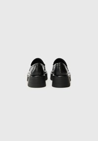 Scarpe in pelle nera con suola piatta spessa, punta arrotondata e texture liscia. La vista posteriore mostra un design minimalista e senza componenti visibili.