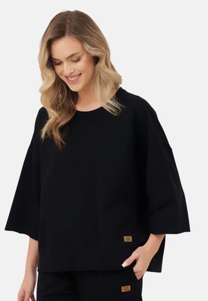 Kobieta o falowanych blond włosach, ubrana w luźną czarną bluzkę z rękawami do łokcia i dopasowane czarne spodnie, uśmiecha się i spogląda w dół z ręką w kieszeni.