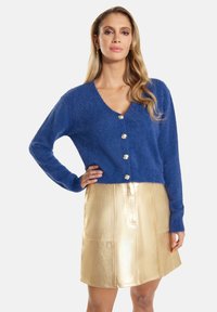 Blauwe fluffy cardigan met een v-hals en zilveren knopen, gecombineerd met een glanzende gouden minirok met voorzakken en gestikt detail.