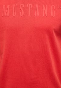 Red cotton t-shirt with embossed lettering "MUSTANG" in matching color. Smooth texture and simple design, featuring a crew neckline.