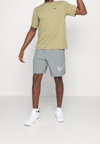 Olivgrön Nike T-shirt, grå Nike-shorts med logotyp, vita skor, med en strukturerad tygtextur synlig i t-shirten och shortsen.
