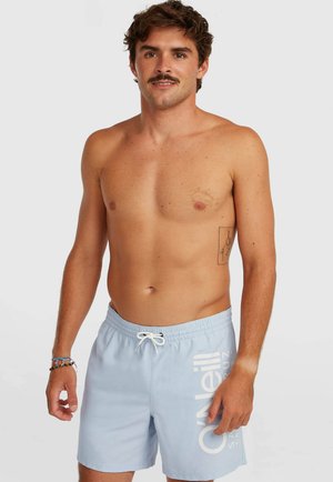 Man met snor die lichtblauwe O’Neill zwembroek en kleurrijke armbanden draagt, staand tegen een effen witte achtergrond.