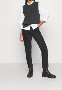 Chaleco de punto gris sobre una camisa blanca, combinado con jeans rectos negros y botas de tobillo negras, contra un fondo neutro.