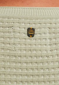 Tejido de punto de color beige claro con un parche rectángulo cosido que presenta una etiqueta negra con la inscripción "PME LEGEND" en texto naranja.