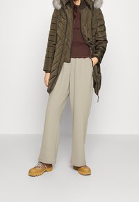 Person in olivgrünem gesteppter Mantel mit Fellkapuze, braunem Rippstrickpullover, beigen weiten Hosen und tannfarbene Wanderstiefel mit dicken Sohlen.