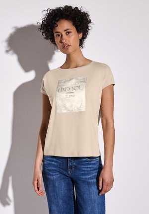 T-shirt beige à manches courtes avec un imprimé graphique carré au centre, comportant du texte. Tissu doux et coupe décontractée. Associé à un jean bleu.