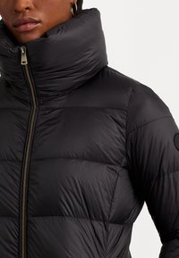 Svart pufferjacka med hög krage, blixtlås och horisontell quiltad design, med en tygpatch på vänster axel.