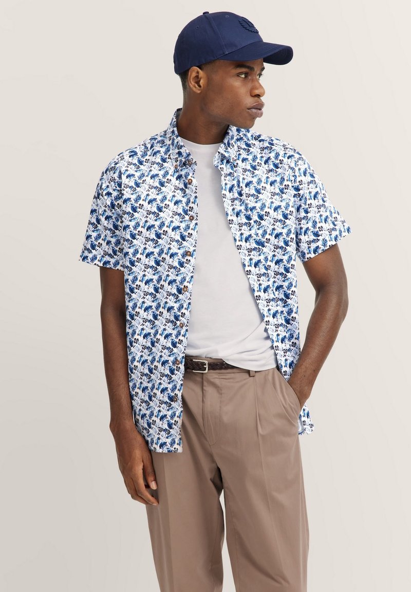 Kurzärmeliges Hemd in Weiß mit blauem Blumenmuster, mit Knopfverschluss, kombiniert mit einem grauen T-Shirt und beigen gefalteten Hosen. Marineblauer Hut.