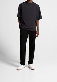 En person står i en enkel svart t-shirt och svarta byxor, iförd vita sneakers mot en enkel, ljus bakgrund.