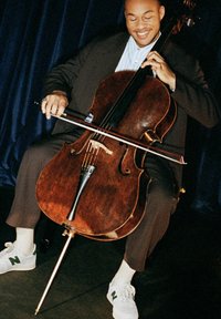 Cello med en mörk träram, böjd form och en glänsande finish. Spelaren håller en stråke, klädd i en mörk outfit och vita sneakers.