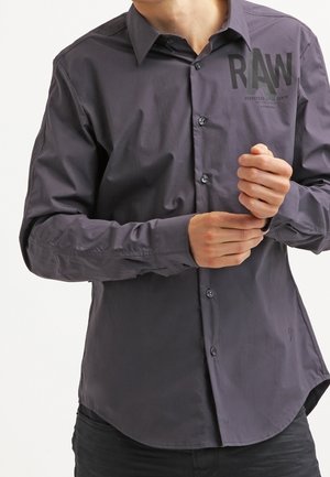 Homme qui ferme le poignet d'une chemise boutonnée gris foncé avec le texte "RAW" sur la partie supérieure gauche de la poitrine.