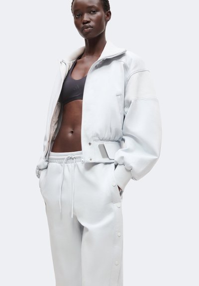 Junge Person trägt eine weiße übergroße Jacke über einem schwarzen Sport-BH und weißen Kordelhosen mit Druckknöpfen an den Seiten.
