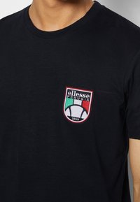 Černé bavlněné tričko s kulatým výstřihem, na kterém je vyšitý štíhový logo v červené, zelené a bílé barvě, zobrazuje fotbal a text "ellesse PERUGIA 1969."
