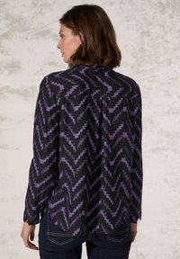Blusa nera con un prominente motivo a zigzag viola, maniche lunghe e un orlo curvo per un tocco in più. Tessuto liscio con una vestibilità comoda.