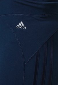 Marinblå träningstights med en slät yta, som har en broderad vit Adidas-logotyp och vertikala velourstripes längs sidorna.