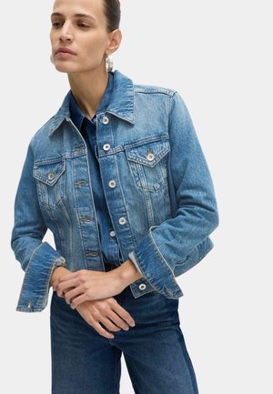 Mujer con el cabello recogido, lleva pendientes de aro plateados, una chaqueta vaquera azul claro con las mangas remangadas sobre una camisa vaquera azul oscura y pantalones vaqueros.