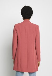 Dlouhý, přiléhavý blejzr v jemně růžové barvě, texturovaná tkanina s vysokým límcem, otevřeným předním dílem a detaily v podobě jediného švu. Spárováno se světle modrými džíny.