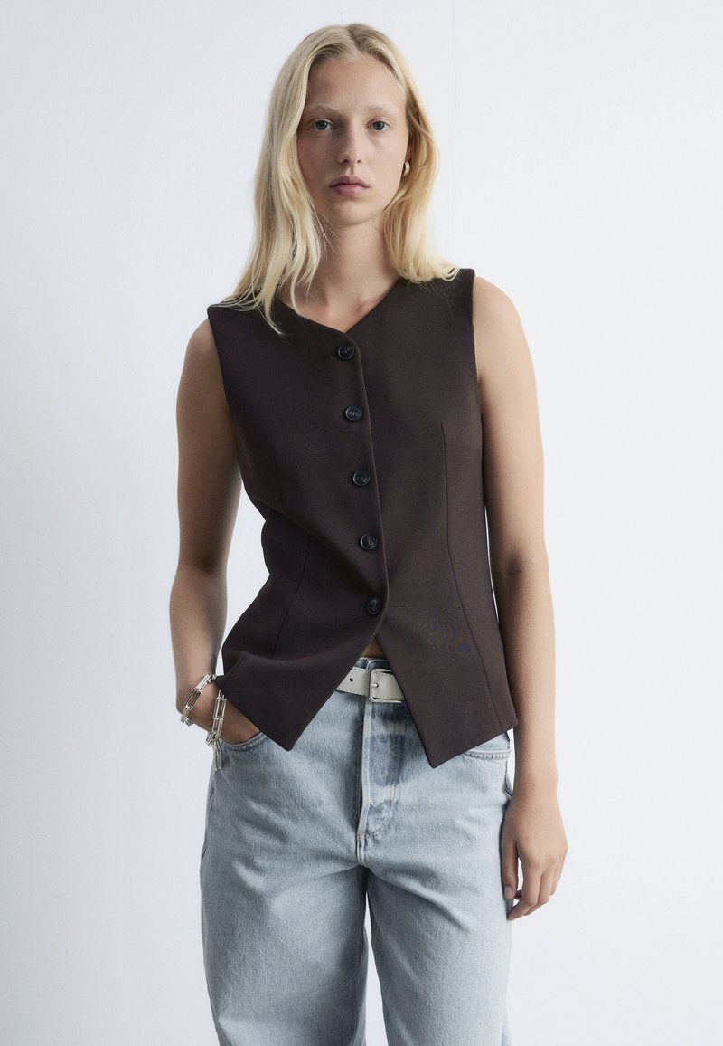 Brauner ärmelloser Blazer mit V-Ausschnitt, ausgestattet mit fünf schwarzen Knöpfen und einer taillierten Passform, kombiniert mit hellblauen hoch geschnittenen Jeans.