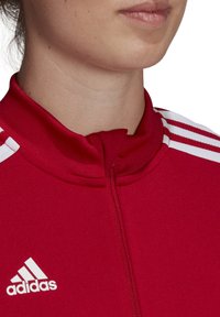Rode zip-up jas met een hoge kraag, met drie witte strepen op de schouders en een wit Adidas-logo aan de linkerkant.