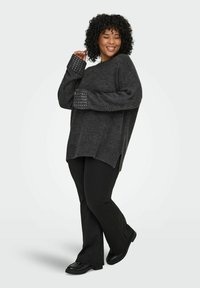 Tamno sivi predimenzionalni pulover s zakovicama na manžetama, bočnim prorezima, nosi se uz crne široke hlače i crne cipele, s teksturiranim pletenjem.