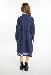 Marineblaues Langarmkleid mit Rüschenausschnitt, Taillennaht und weißem Zickzackbesatz am Saum und den Manschetten. Getragen mit schwarzen Knöchelstiefeln.