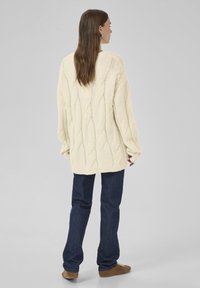 Frau trägt einen übergroßen, cremefarbenen Kabelstrickpullover, dunkelblaue Jeans und braune Sneaker, steht mit dem Rücken zu einem schlichten grauen Hintergrund.