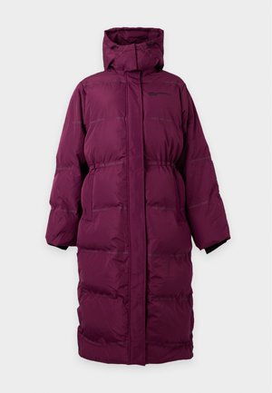 Femme portant un long manteau matelassé bordeaux et un bonnet assorti, un haut et un pantalon noirs, ainsi que des chaussures noires et blanches, debout devant un fond uni.