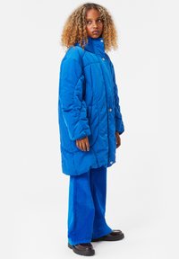 Blå, överdimensionerad puffjacka med hög krage och knäppning; matchad med blå kunnebyxor och svarta klumpiga skor.