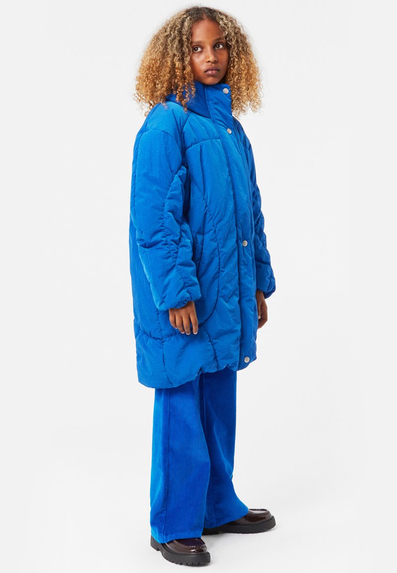 Blå, överdimensionerad puffjacka med hög krage och knäppning; matchad med blå kunnebyxor och svarta klumpiga skor.