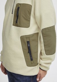 Fleece pullover in cream with green fabric patches. Features a quarter zip and multiple zippered pockets. Soft texture and casual fit.