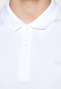 Polo blanc en tissu texturé, doté d'une patte de boutonnage à trois boutons et d'un petit logo bleu clair sur le côté gauche.