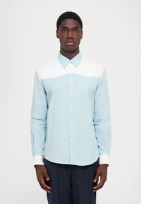 Camicia azzurro chiaro con yoke e polsini bianchi, dotata di colletto, due tasche frontali e una texture liscia.