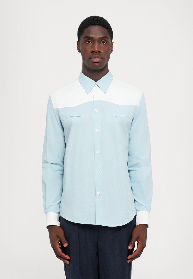 Camicia azzurra a maniche lunghe con yoke bianco, apertura con bottoni, e due tasche sul petto. Presenta un colletto a punta e polsini bianchi a contrasto.