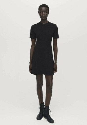 Woman standing wearing a fitted short-sleeve black dress and black lace-up ankle boots against a plain light background.