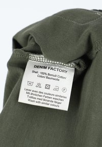 Olivgrön bomullstyg med en skötselätt som visar tvättanvisningar och varumärket "DENIM FACTORY" på flera språk.