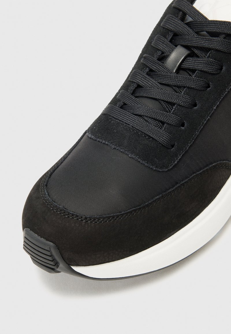 Chaussure de sport noire composée d'une combinaison de textile lisse et de suède, à bout rond, avec des lacets noirs texturés et une semelle blanche.