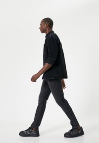 Camicia di denim nera con maniche arrotolate, abbinata a jeans grigio scuro e sneakers nere con un motivo testurizzato. Modello che cammina.