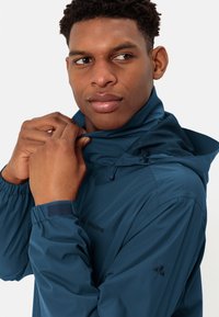 Blå vattentät jacka med hög krage, justerbar huva, elastiska ärmslut och logotypdetaljer; slät textur och lätt design.