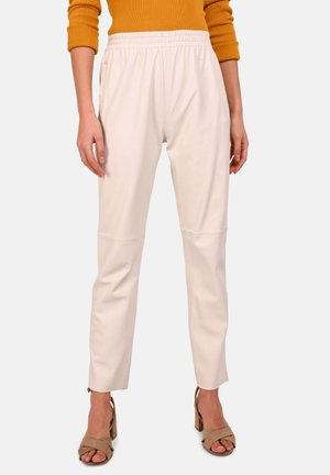 Vrouw die beige rechte broek met hoge taille en elastische tailleband draagt, gecombineerd met tankleurige sandalen met hak en een oranje top met lange mouwen.