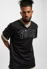 Tricou din plasă neagră cu detalii bej contrastante și logo-ul "FUBU" pe față. Are mâneci scurte și un design cu decolteu în V.