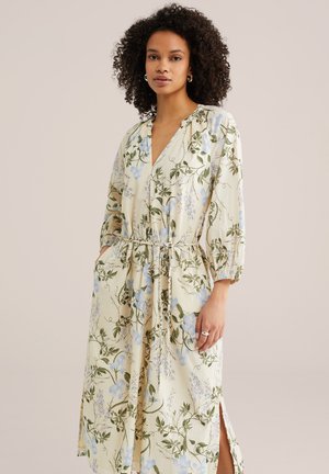 Kobieta z kręconymi włosami, ubrana w długą kremową sukienkę w niebieskie kwiaty, z rękawami o długości do łokcia, dekoltem w szpic i wiązaniem w talii, z rękami w kieszeniach.