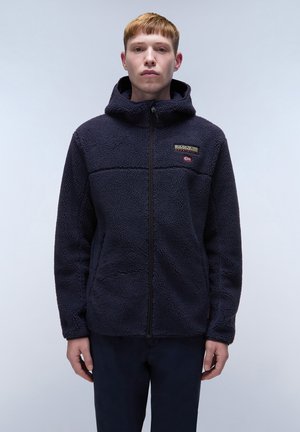 Junger Mann trägt eine marineblaue Fleecejacke mit Reißverschluss und Kapuze und steht vor einem schlichten, hellen Hintergrund.