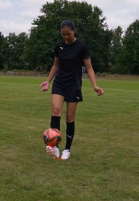 Svart Puma sportskjorte, matchende shorts og svarte sokker. En spiller balanserer en oransje og svart fotball på foten, med grønt gress som bakgrunn.