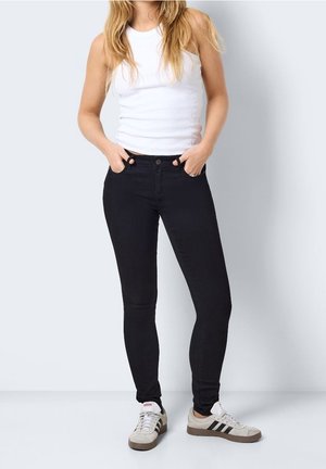 Kvinde iført en hvid ærmeløs top, sorte skinny jeans og hvide Adidas-sneakers med sorte striber, stående med hænderne i lommerne.