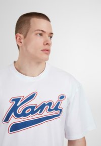 White cotton t-shirt featuring a blue cursive "Kani" logo with red accents. The design has a crew neckline and short sleeves.