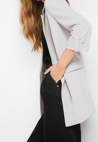 Blazer gris clair avec des manches retroussées, détail à un bouton. Associé à un haut et un pantalon noirs, mettant en valeur une coupe décontractée.