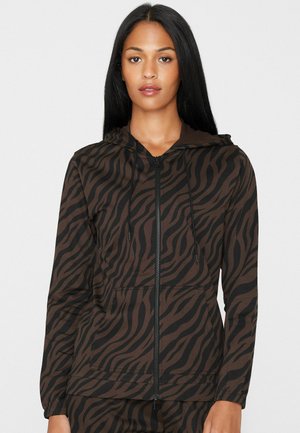 Mujer con cabello largo y negro que lleva una sudadera con capucha con cremallera de rayas de cebra marrón y negro, con bolsillos delanteros y capucha.