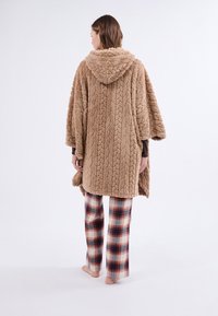 Hnědý texturovaný pončo s kapucí a vzorem z copánkového pletení; nošeno přes kostkované pyžamové kalhoty v krémové, červené a námořnické barvě; měkký fleecový materiál.